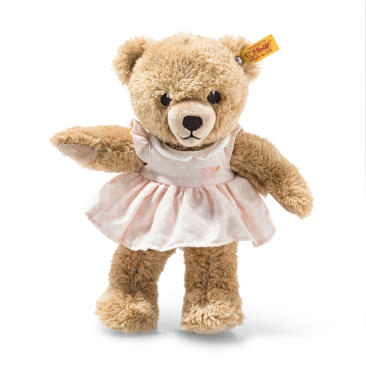 This is Sleep Well Bear by Steiff. This medium bear has beige fur that is long, slightly shaggy with soft curls and a low sheen. The muzzle and paw pads are a lighter white tone that contrasts with the beige fur. Inner ears match the beige fur. Eyes are black and round. The nose is black and rounded with an embroidered black mouth below. There are no distinct markings beyond uniform fur. The bear wears a removable pink dress with white polka dots and a small embroidered motif on the bodice; the dress h...