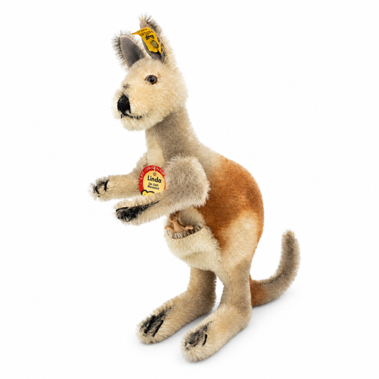 This is Linda by Steiff. Standing handmade kangaroo made of mohair with a firm feel, shown upright on its hind legs. Fur is beige across head, chest, limbs and pouch, with warm brown on the back and rump and a slightly gray tail; short, fine, slightly shaggy fur with a low sheen. Muzzle is beige, inner ears are darker gray, paw tips and small claw markings are black, and there are black markings around the mouth and nose; eyes are brown and the nose is black and rounded. A small plastic joey sits in th...