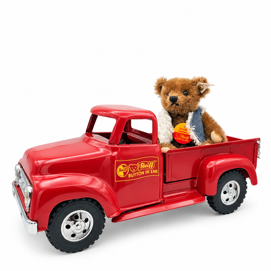 This is Special Delivery by Steiff. Small sitting bear made of mohair with short to medium, slightly shaggy fur that has a soft sheen. Fur is brown overall with a beige muzzle, beige inner ears, beige paw fronts and beige paw pads; the chest/tummy area appears the same brown as the rest of the fur. Eyes are black and round; nose is black and triangular. Bear wears a blue denim vest with a white sherpa-style trimmed edge; the bear is seated in the bed of a red pickup truck accessory. The bear is fully j...